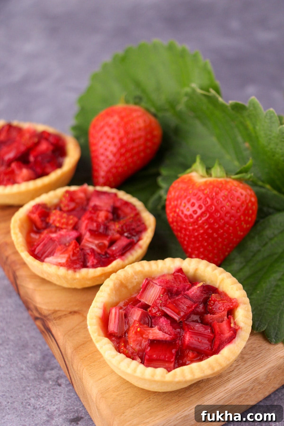 Row of strawberry rhubarb tartlets on a chopping board, with fresh strawberries and strawberry leaves on the side.