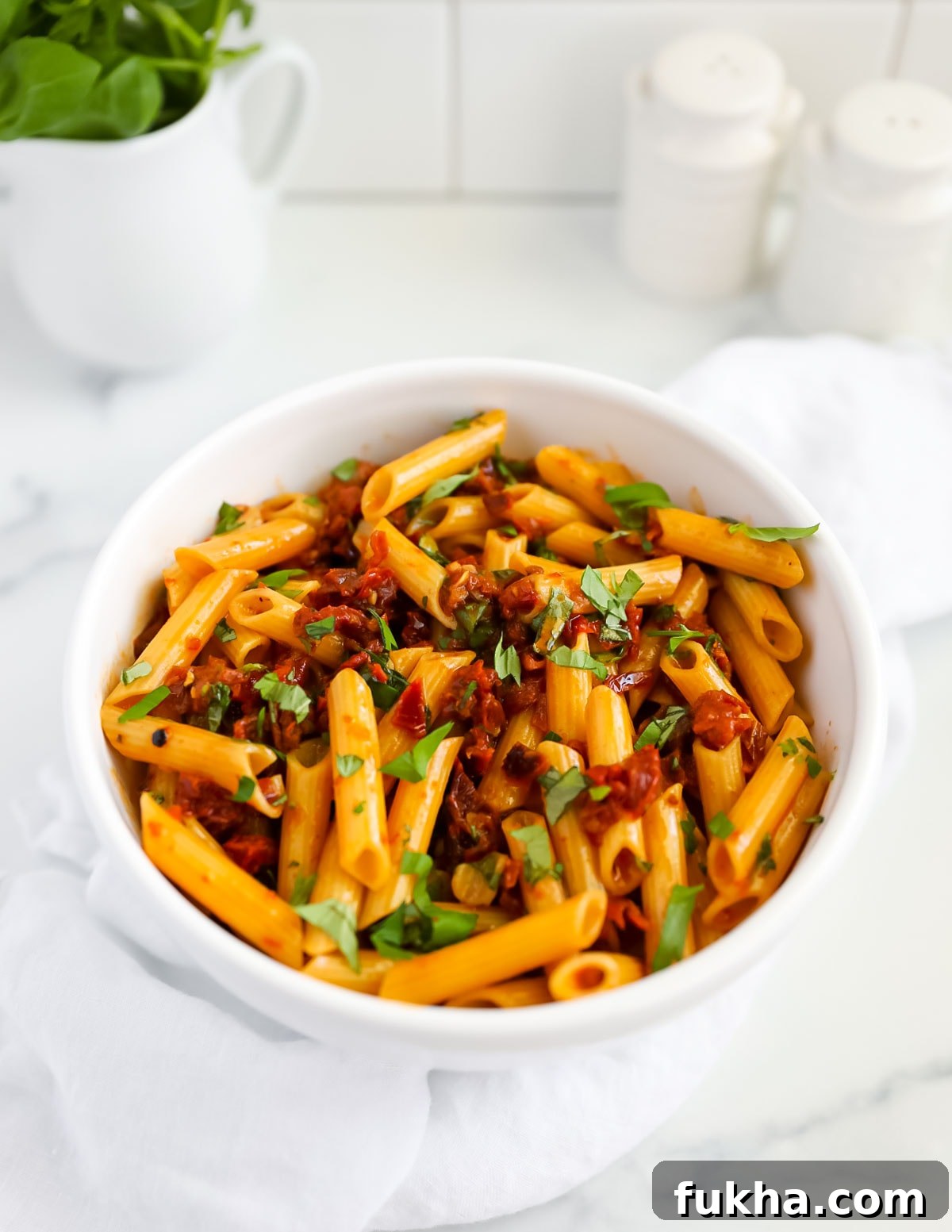Sun Dried Tomato Pasta Recipe with Garlic and Basil 2 A white bowl filled with penne pasta and chunky red pasta, covered in chopped fresh green basil leaves