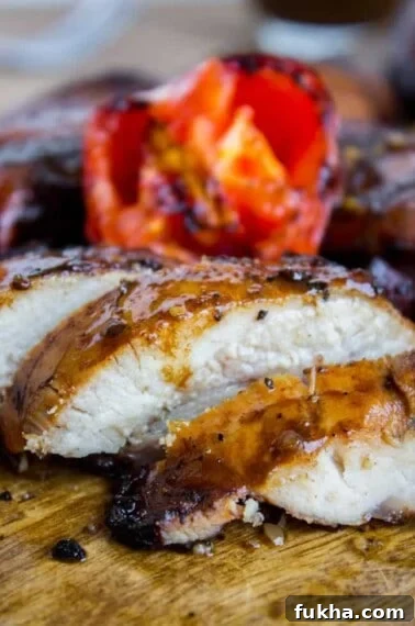 Close-up of juicy grilled chicken breast sliced on a wooden board, with a charred tomato in the background.