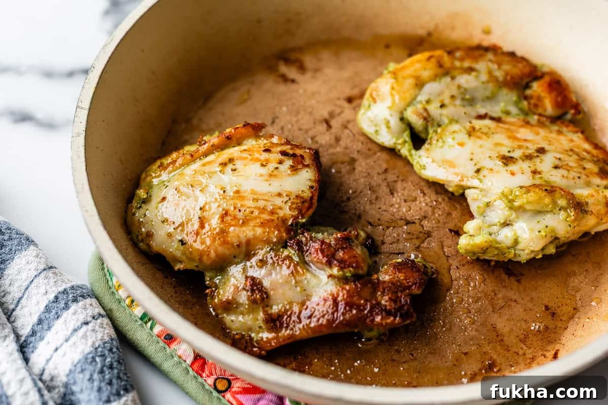 Two browned chicken thighs are cooking in a beige skillet with some oil, resting on a patterned oven mitt. A striped kitchen towel is visible nearby on a marble countertop.