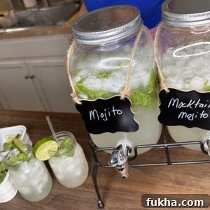 Two large glass dispensers labeled “Mojito” and “Mocktail Mojito” sit on a counter, filled with ice, lime, and mint. In front, two mason jars with drinks, lime slices, and a tray with limes and mint are visible.