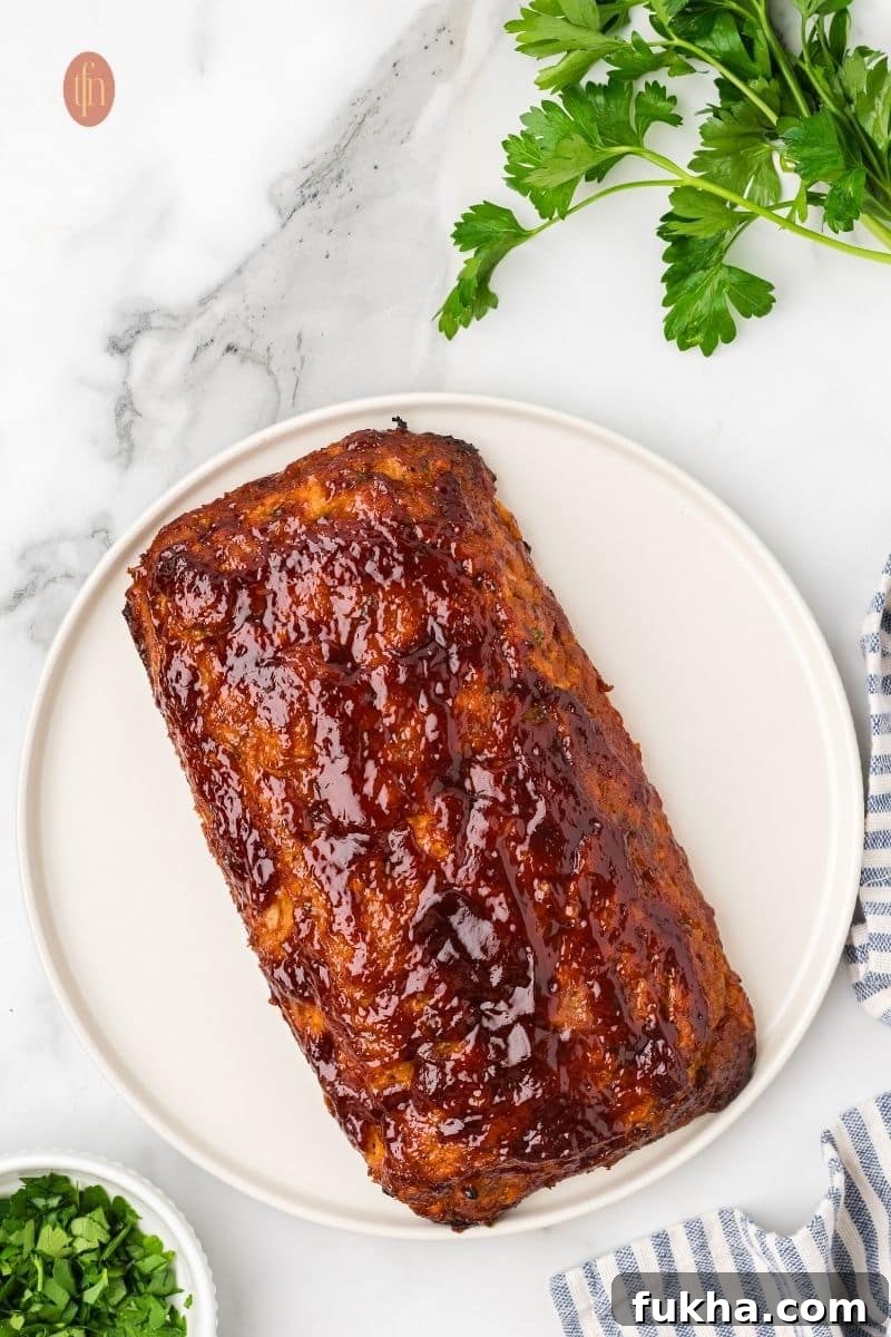 An overhead shot of a whole chicken meatloaf on a white platter. It is covered in a glossy, dark red glaze. A sprig of parsley and a blue and white striped towel are nearby.