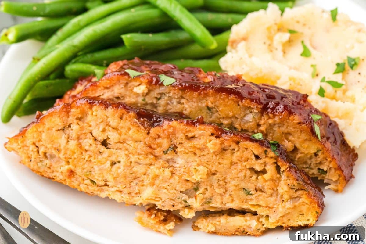 A wide, close-up shot of two slices of glazed chicken meatloaf served on a white plate with green beans and mashed potatoes.