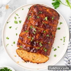 A square, overhead shot of a whole chicken meatloaf on a round white platter. It is already sliced and topped with a glossy glaze and chopped parsley.