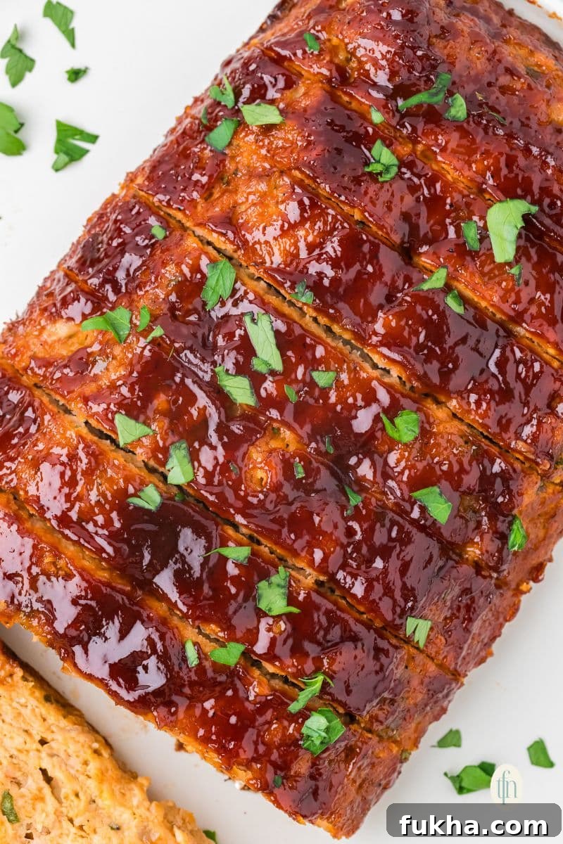 A close-up, overhead view of a sliced chicken meatloaf. The top is covered in a thick, shiny red glaze and sprinkled with chopped parsley.
