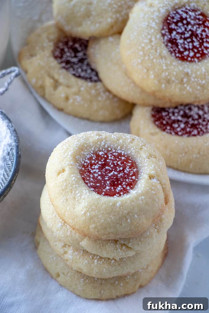 Classic, easy and delicious with only 7 ingredients these Thumbprint Cookies the perfect quick and tasty cookie recipe to bake up for the holidays! Thumbprint Cookies stacked on top of one another with plate of cookies in background
