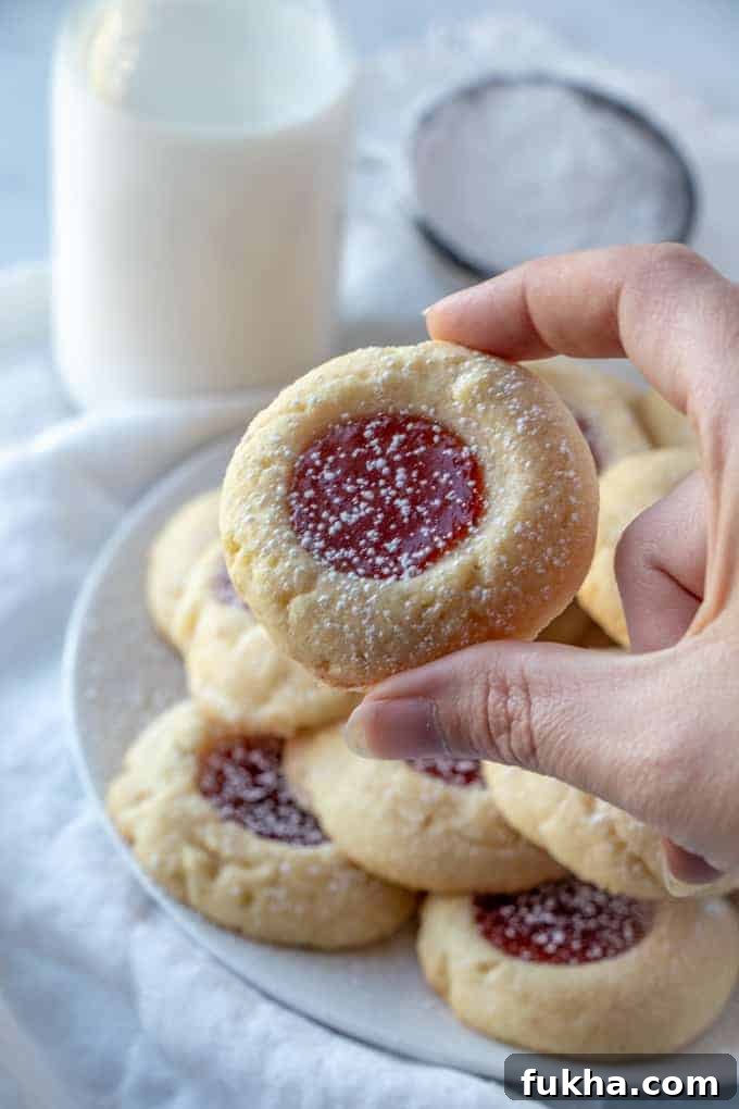 Classic, easy and delicious with only 7 ingredients these Thumbprint Cookies the perfect quick and tasty cookie recipe to bake up for the holidays! hand holding one thumbprint cookies with plate of cookies and milk in background